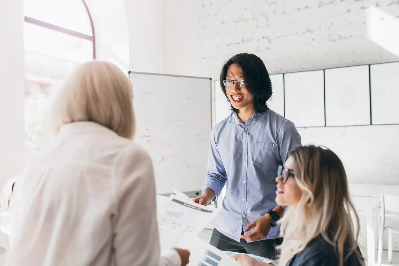 Rückenansicht einer blonden Frau in einem stylischen Hemd, die Dokumente liest, während ein asiatischer Mann ihr etwas sagt.